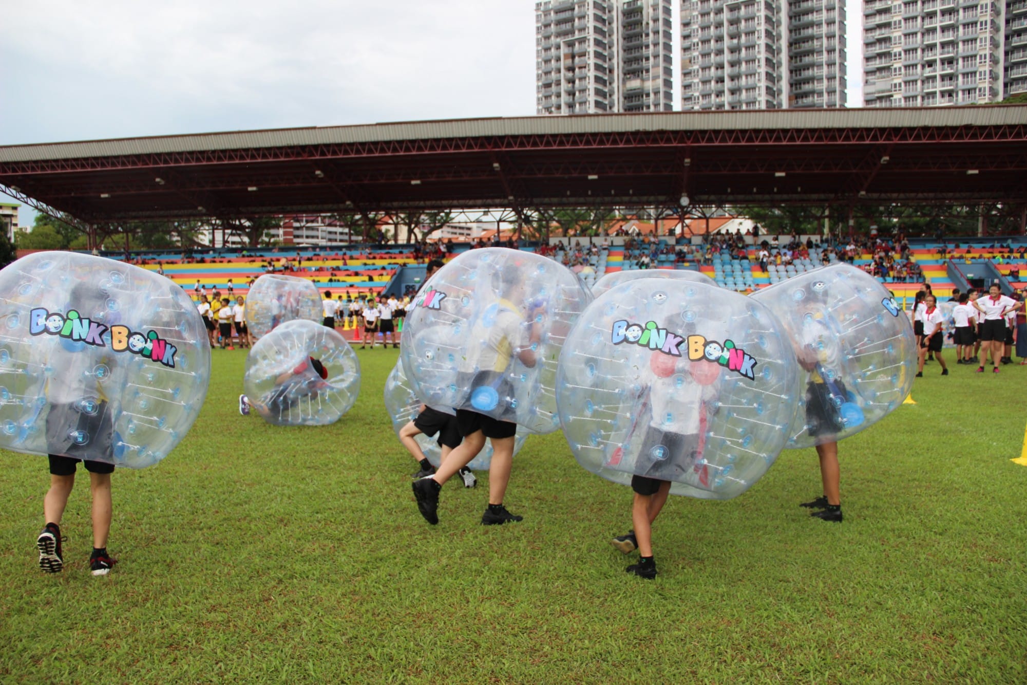 Children wearing large transparent inflatable bubble suits play on a grass field at a stadium.